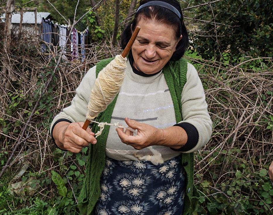 Older Albanian woman holding yarn wrapped around a stick in front of brambles