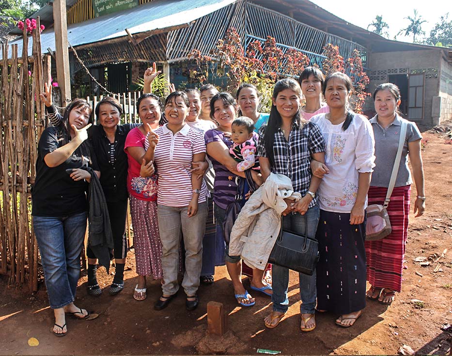 A group of Thai women stand in front of a fence and in front of a building made from sticks