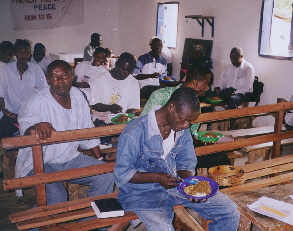 Liberian men sit on rows of wooden benches eating lunch