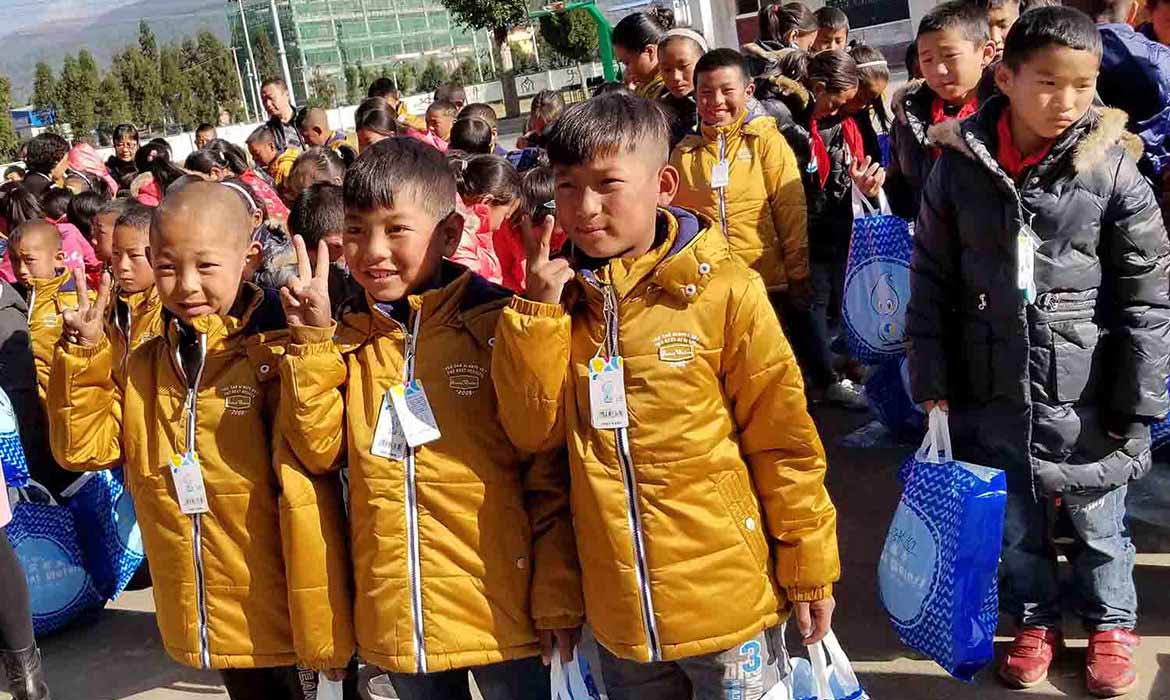 Three Chinese boys in yellow coats stand in front of a group of Chinese kids holding up a one or two with their fingers