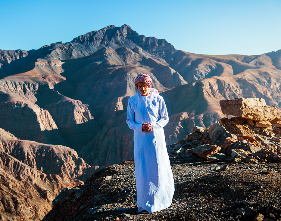 Emiratis man standing on the top of a rocky mountain with other rocky mountains behind him.