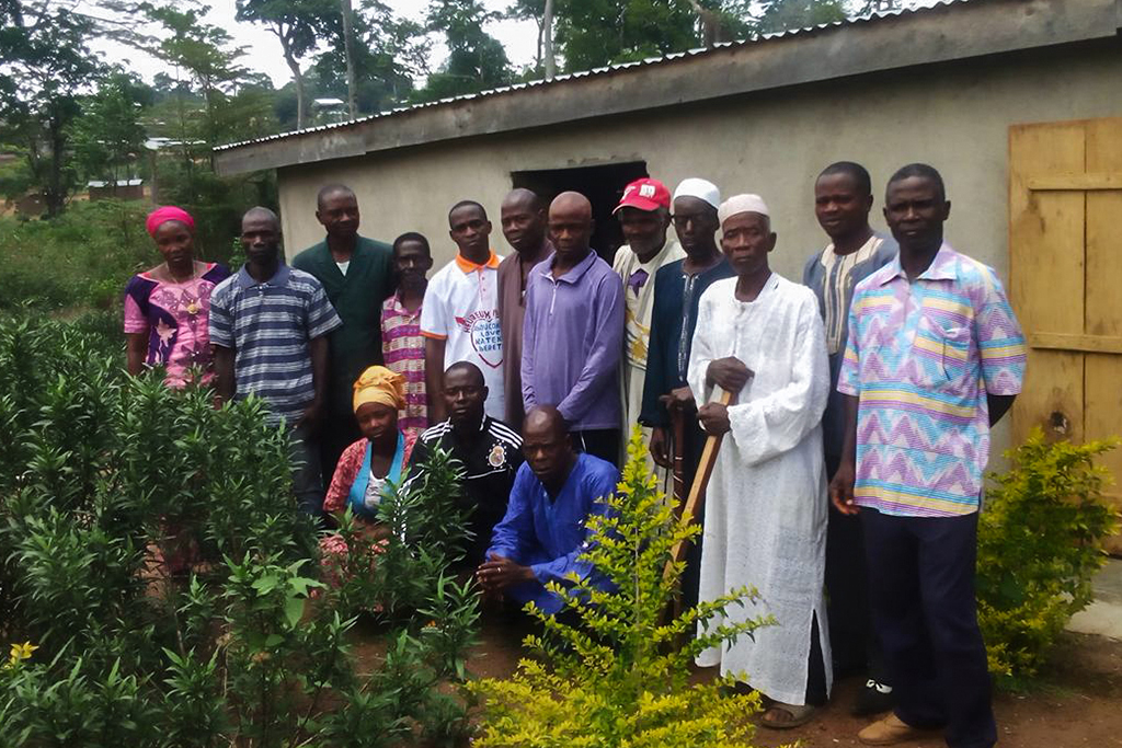 Ivorian Christian men and women standing behind bushes in front of a concrete house