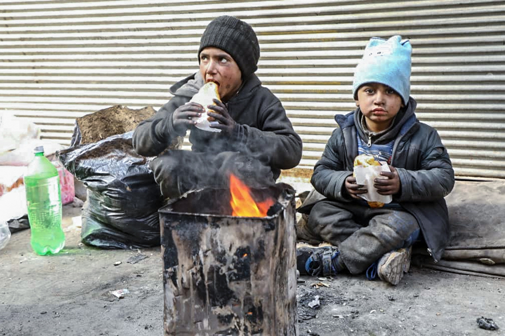Two Syrian boys covered in soot sit on a dirty street eating sandwiches with a fire burning in front of them for warmth