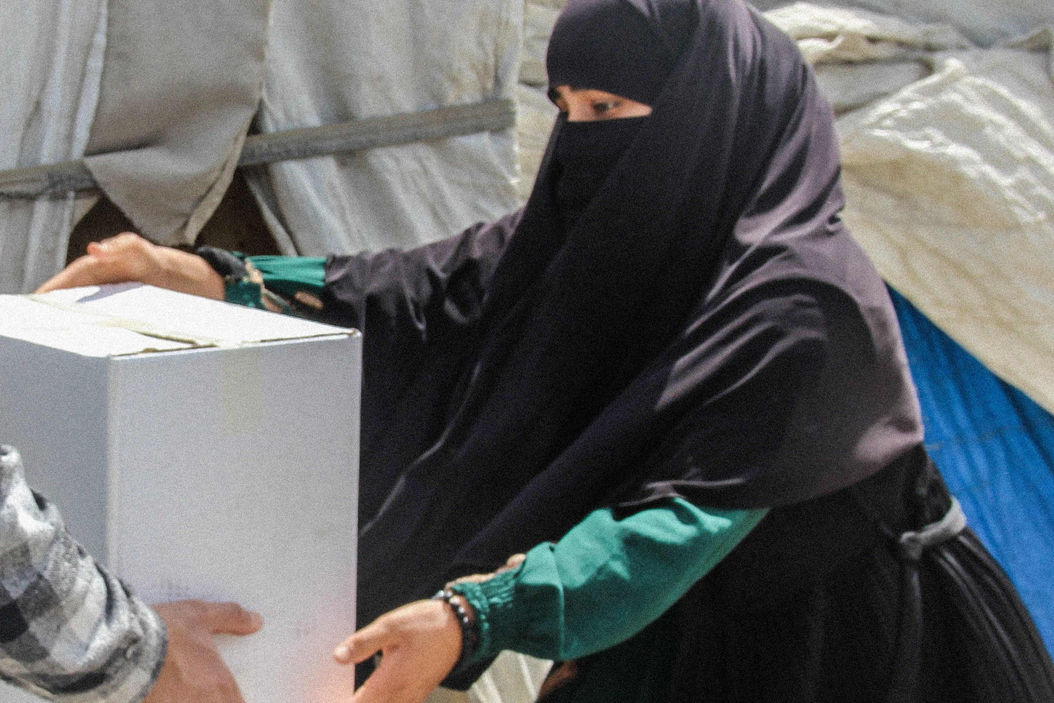 A woman wearing a hijab accepts a box of aid from a worker in a refugee camp