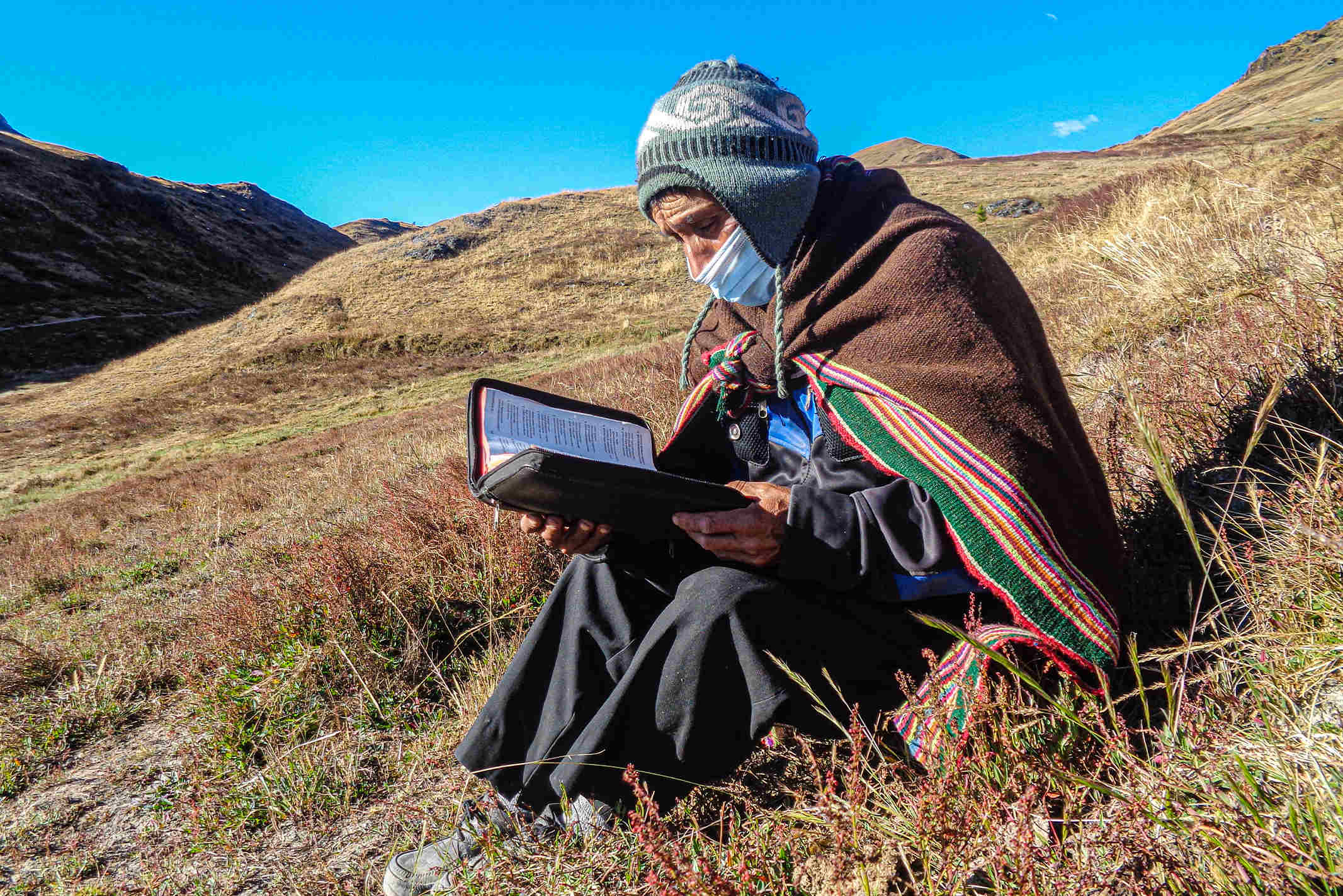 A Peruvian Christian missionary sitting in a grassy valley wearing a shaw reading his Bible