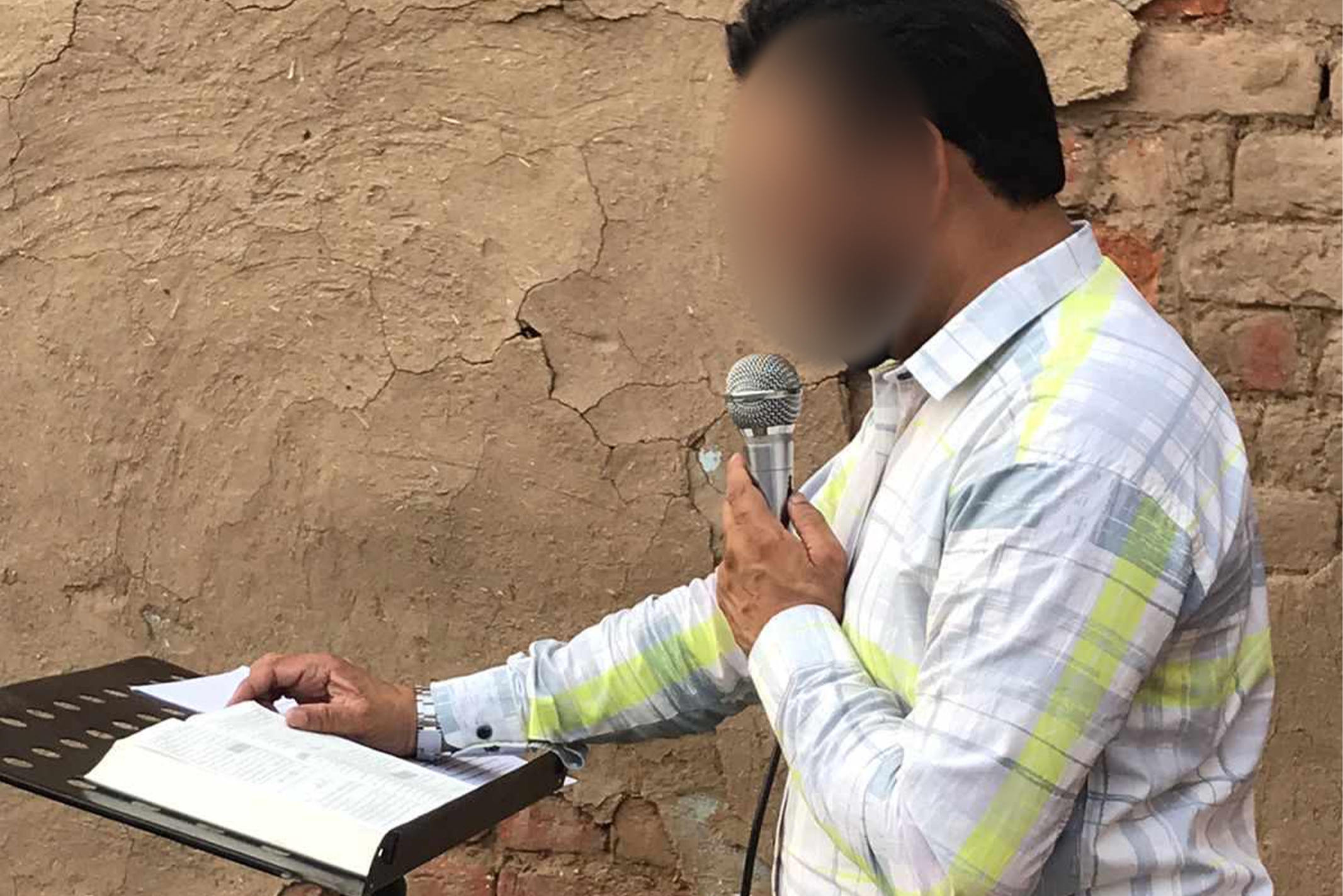 Pakistan Christian pastor preacher while holding microphone and standing next to a brick wall